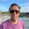 Headshot of a man wearing sunglasses, stood on a beach with blue sky behind him