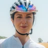 Head shot of cyclist Manon Lloyd wearing a white cycling helet, rainbow sunglasses and a white cycling jersey.
