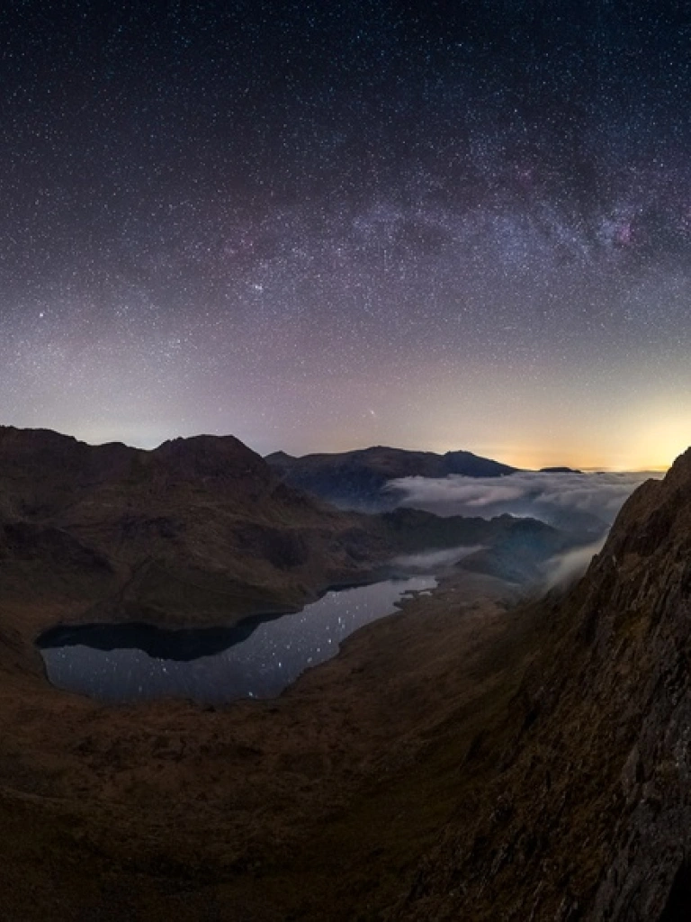 Copaon mynyddoedd Eryri yn erbyn awyr dywyll gyda sêr a golau oren machlud haul ar y gorwel.