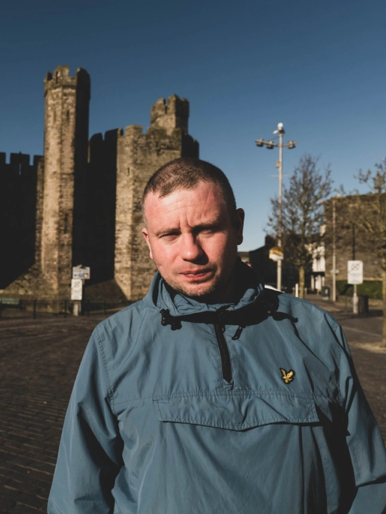 Person mewn cot las yn sefyll o flaen Castell Caernarfon. 