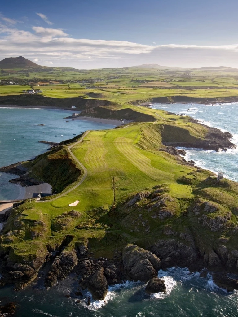 Golygfa o'r awyr o gwrs golff Nefyn ar benrhyn yn ymestyn tua'r tonnau.