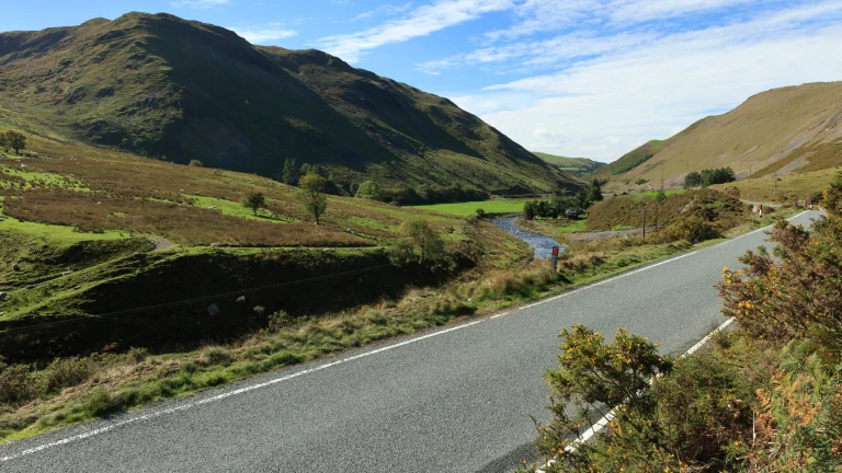 Llun o ffordd gyda bryniau, nant a chaeau yn y cefndir
