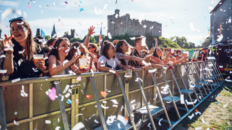 Cynulleidfa gig cerddoriaeth yn Tafwyl yn sefyll tu nôl i wahanfur gyda chonffeti o'u cwmpas a Chastell Caerdydd yn y cefndir