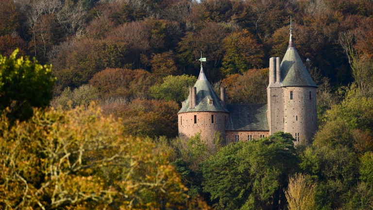 Dau dŵr castell cerrig yn codi uwchben y coed yn yr hydref ar ochr bryn coediog.
