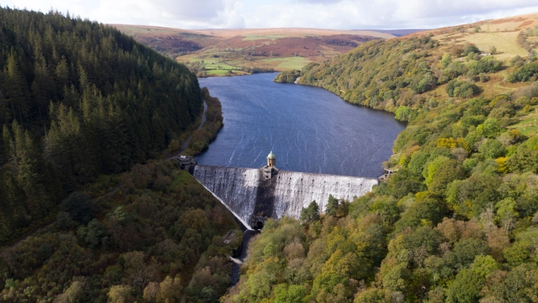 Golygfa o'r awyr o afon yn llifo dros argae agored gyda coed a mynyddoedd o gwmpas