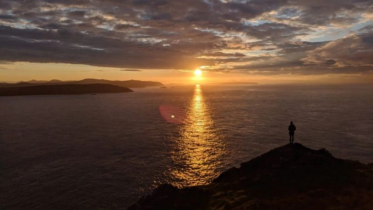 Person yn sefyll ar ymyl clogwyn yn edrych allan tuag at y môr, yn gwylio'r machlud