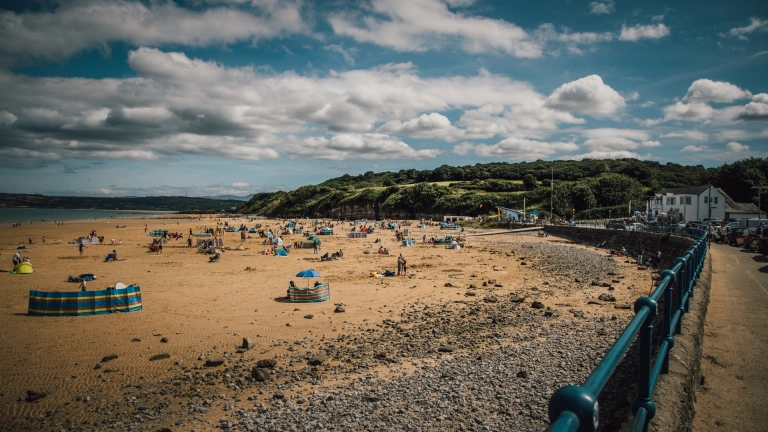 Traeth prysur tywodlyd. Mae'r awyr yn las ond yn gymylog.