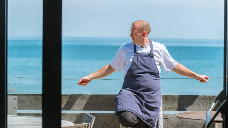 Y pen-cogydd Hywel Griffith yn ei fwyty Beach House gyda'r môr yn y cefndir.
