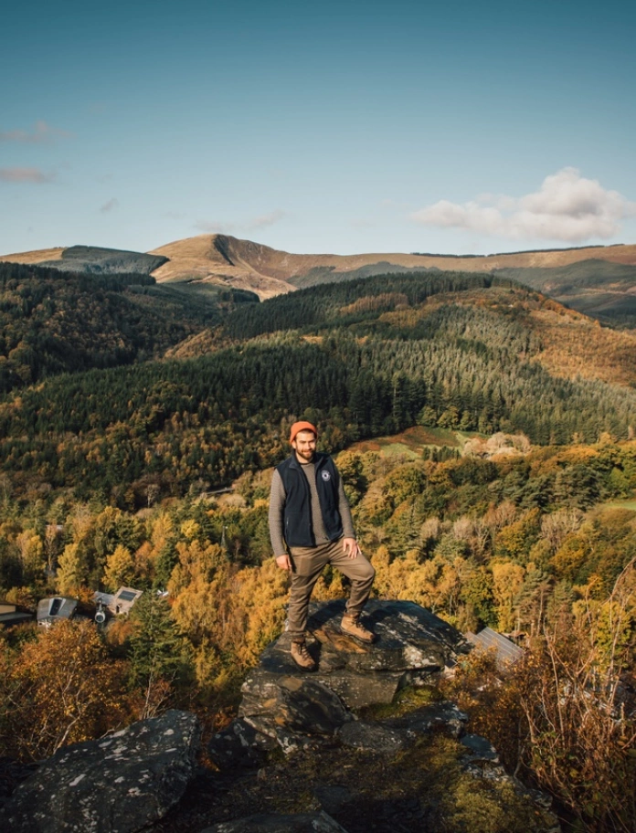 Dyn yn sefyll ar gefnen gyda thirwedd y tu cefn iddo.