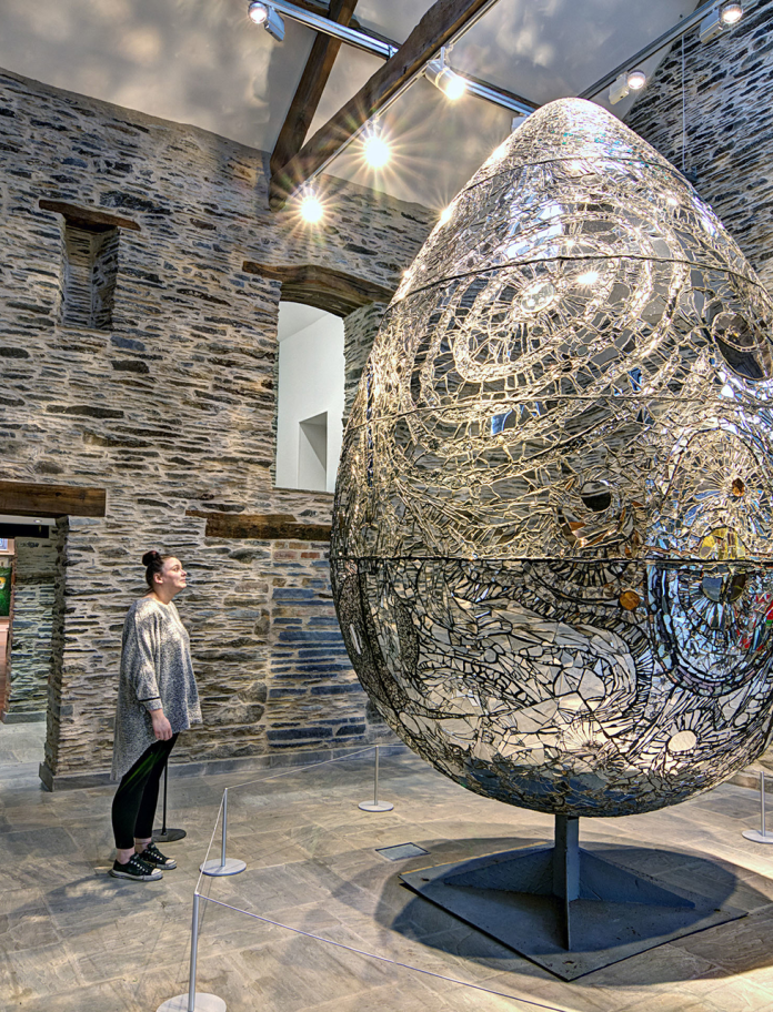 Cerflun wy anferth gyda person yn sefyll yn edrych arno y tu mewn i ystafell wal gerrig.