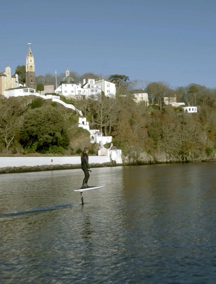 Dau berson yn defnyddio byrddau syrffio eFoil ar yr afon o flaen Portmeirion.