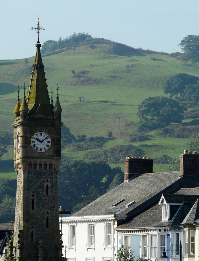 Twr cloc Machynlleth gyda thai wrth ei ochr a bryniau gwyrdd tu cefn.