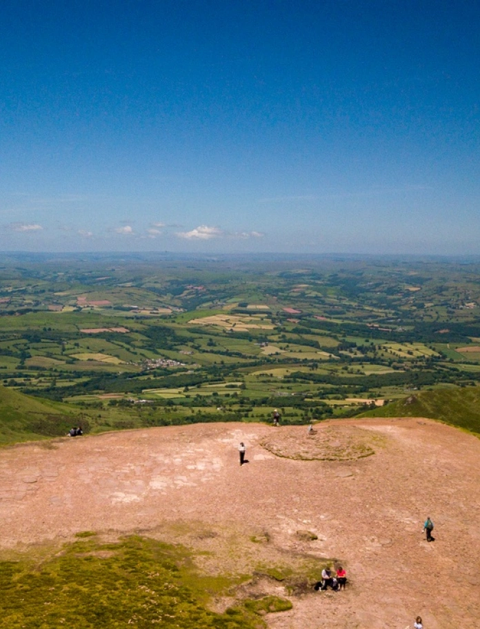 Golygfa o'r awyr o'r gwastadeddau gwyrdd islaw o ben Pen y Fan gydag awyr las.