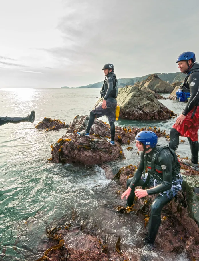 group of male adults laughing as they jump off the rocks into the blue sea, coasteering with instructors.