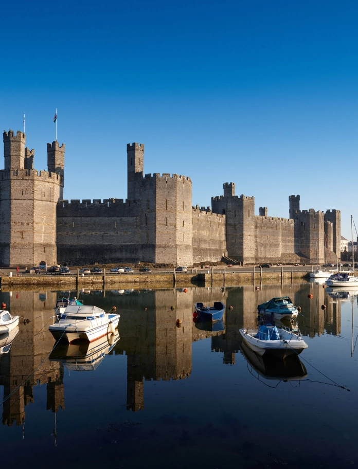 Llun o Gastell Caernarfon o'r de, gyda chychod yn yr harbwr o flaen y castell