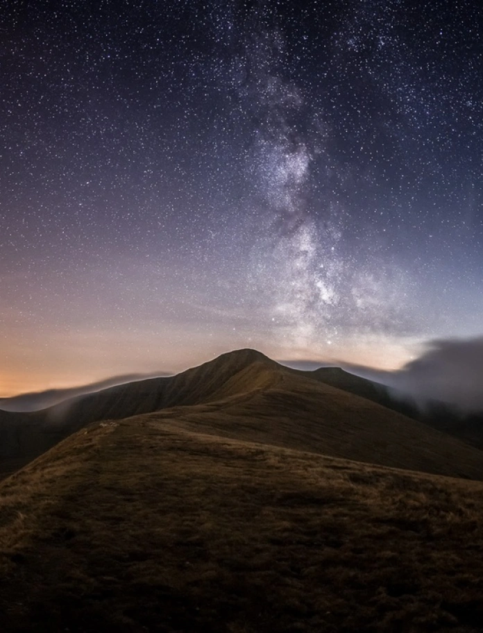 Awyr dywyll yn llawn sêr uwchben tirwedd fynyddig Bannau Brycheiniog gyda golau'r haul yn machlud ar y gorwel.