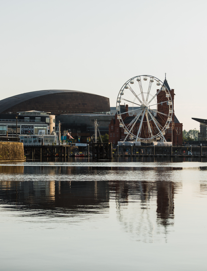 Golygfa o Fae Caerdydd o’r dŵr sy’n dangos adeiladau ac olwyn fawr