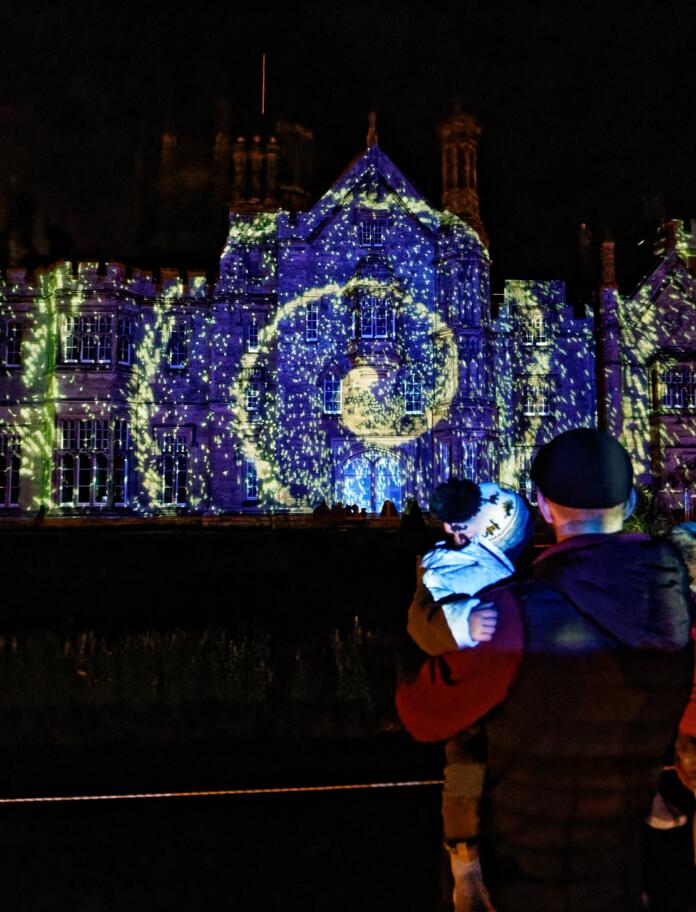 man and woman holding children watch light show projected onto manor house.