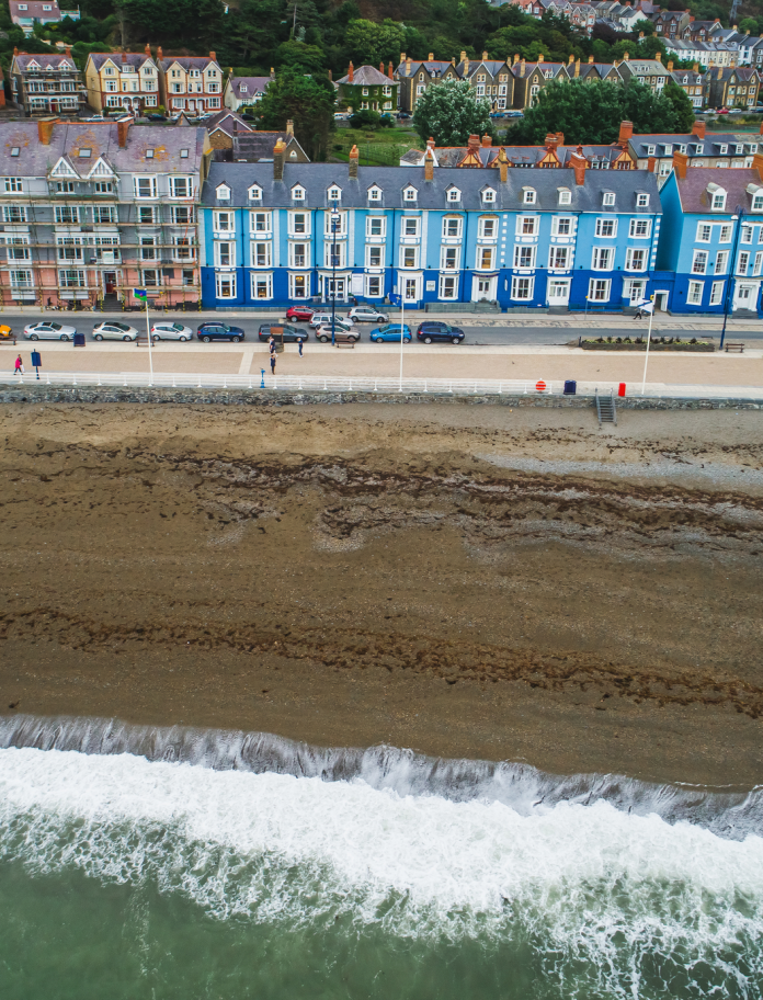 Golygfa o dai o wahanol liwiau’n edrych dros draeth tywodlyd a môr.