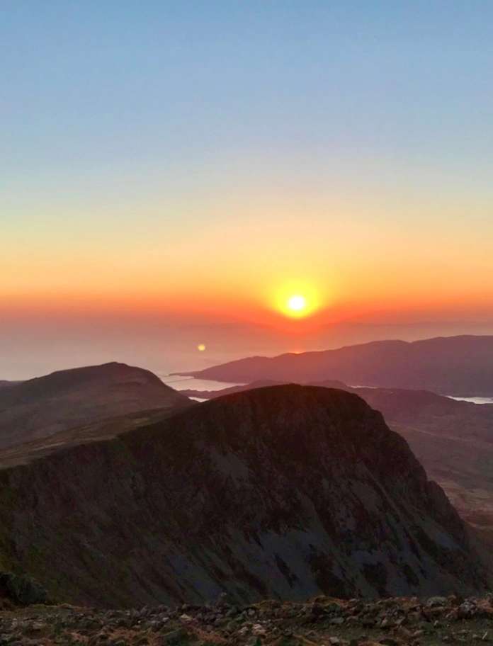 Machlud haul o uwch ben mynydd (Cader Idris). Mae'r awyr yn oren a las ac mae'r mynyddoedd ym mlaen y llun yn wyrdd tywyll.