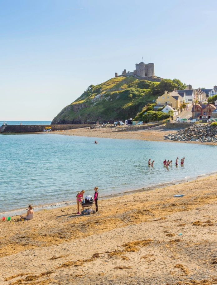 Lan y môr Cricieth gyda thywod euraidd a phobl yn mwynhau ar y traeth. Mae'r môr a'r awyr yn las a gellir gweld Castell Cricieth ar graig yn y pellter.