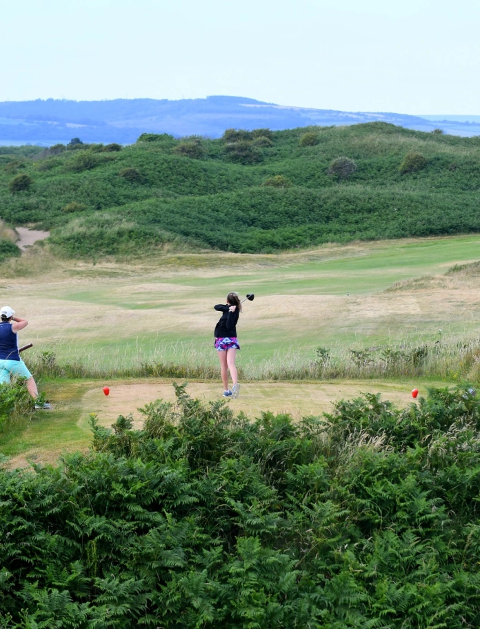 Golffwraig yn taro’r bêl ar gwrs golff yng nghanol twyni glaswelltog.