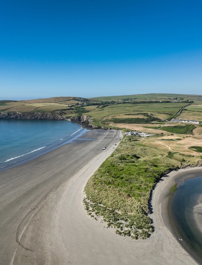 Golygfa o'r awyr dros gwrs golff wrth ymyl traeth tywodlyd.