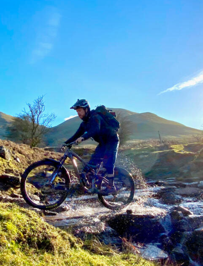 Dyn ar feic mynydd yn mynd drwy nant.
