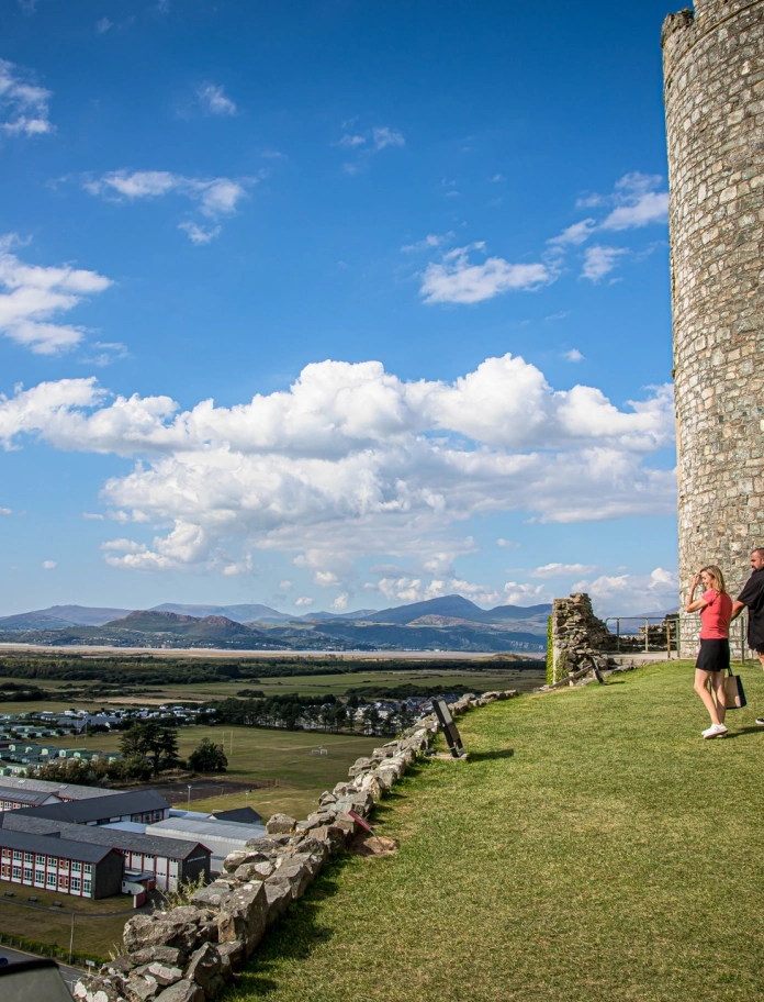 Dau berson yn cerdded ger y castell gyda golygfeydd tua'r mor a'r mynyddoedd.