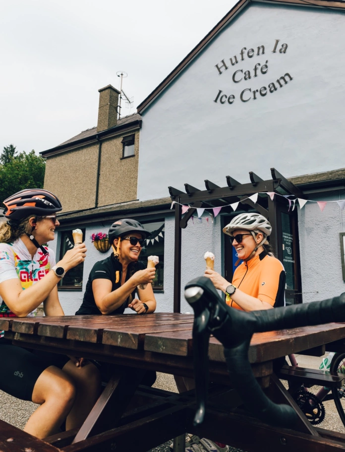three people with cycling helmets sat on bench with ice cream cones and a bike.
