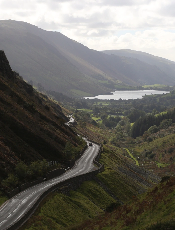 Car yn gyrru i fwrdd o'r camera ar hyd ffordd fynyddig, droellog, gyda llyn a mynyddoeddd yn y cefndir