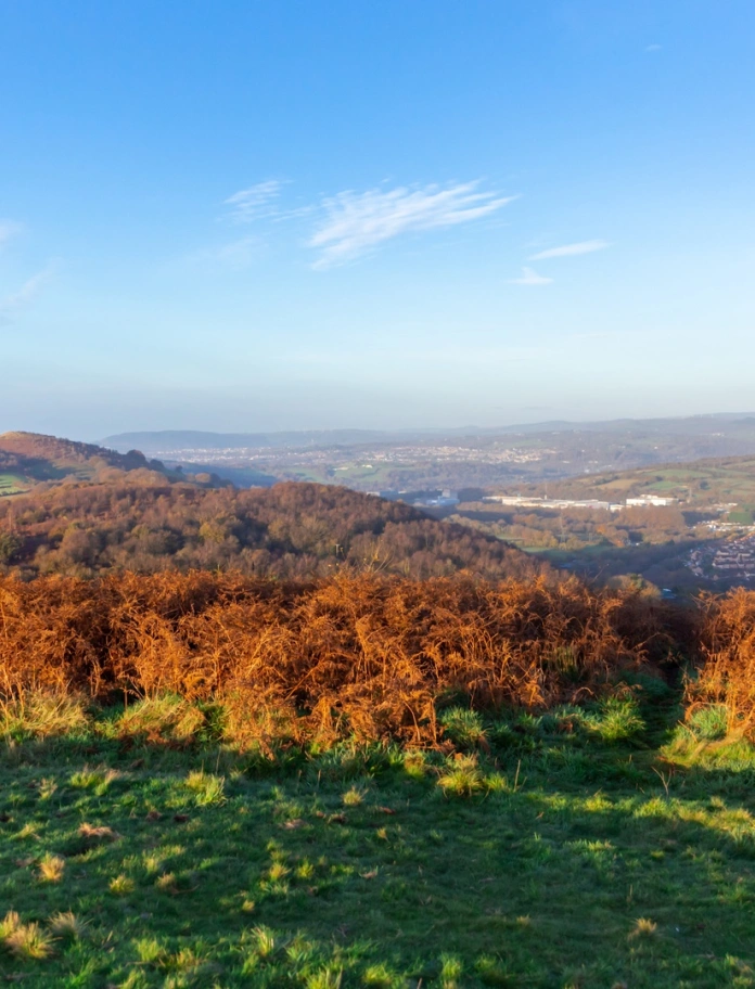 golygfeydd o gopa mynydd, yn edrych dros dref, gyda bryniau gwyrdd o’i hamgylch.