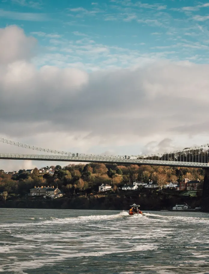 Llun o'r bont grog gyda choed a thai yn y cefndir a chwch ar Afon Menai