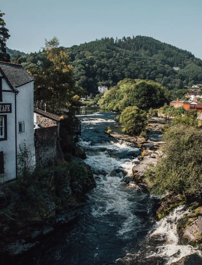 Afon yn llifo ger adeilad gwesty mawr gwyn mewn tref. Mae'r awyr yn las ac mae bryn o goed yn y cefndir.