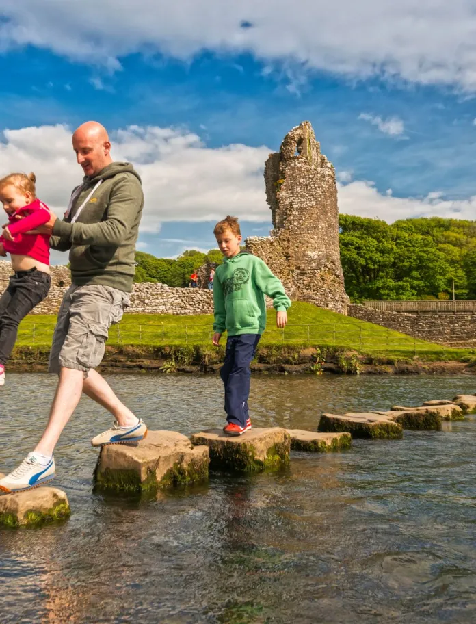Teulu yn croesi Afon Ogwr ar gerrig o flaen y castell