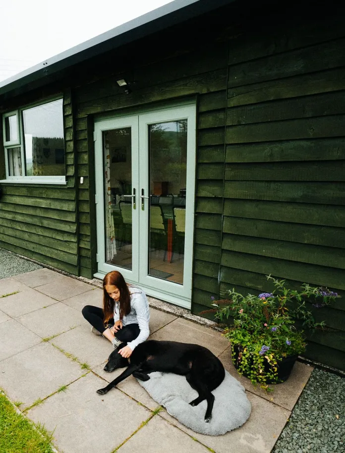 Merch a ci yn eistedd ar batio tu allan i adeilad steil chalet yn y wlad.