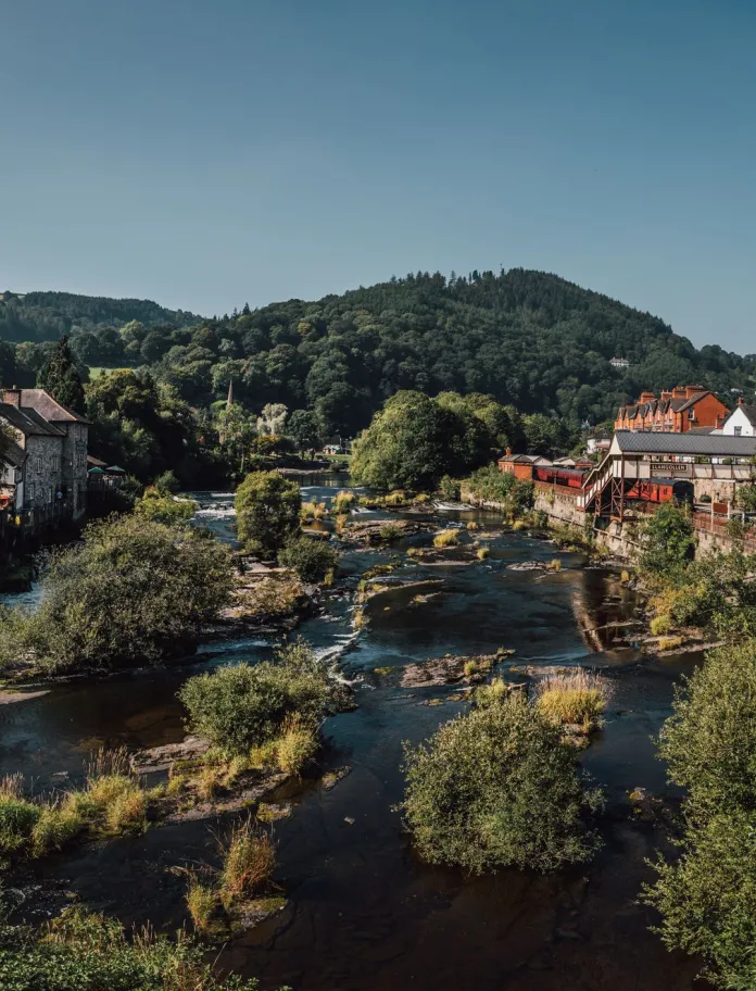 Afon lydan, fas yn rhedeg drwy dref, gyda gorsaf drenau ar un ochr ac adeiladau ar yr ochr arall.