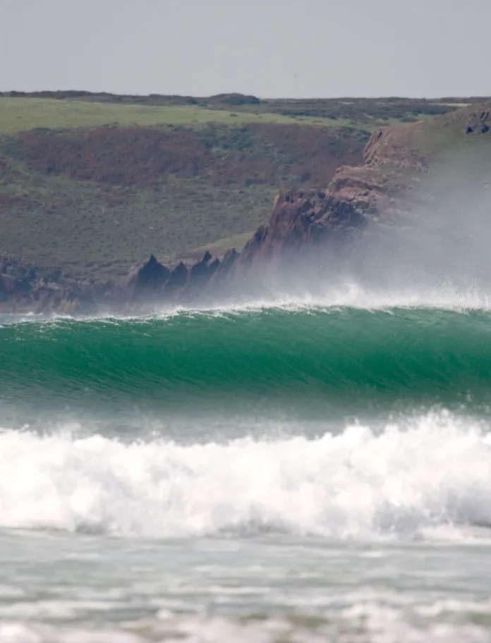 Ton yn torri yn y môr ger y lan yn Freshwater West, Sir Benfro, Gorllewin Cymru, gyda chlogwyni a bryniau glaswelltog yn y cefndir.