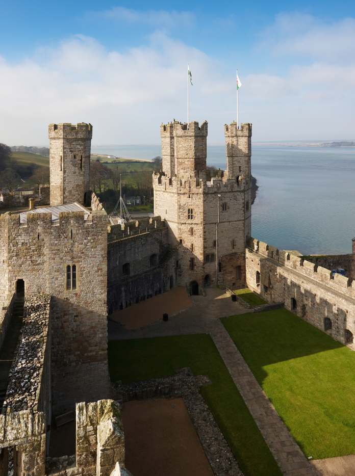 Golygfa o du fewn i Gastell Caernarfon, gyda gwair a thyrrau yn y tu blaen, a'r môr yn y cefndir 