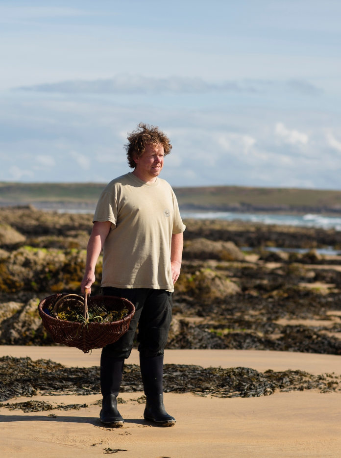 Y chwilotwr Matt Powell yn sefyll ar y traeth yn dal basged