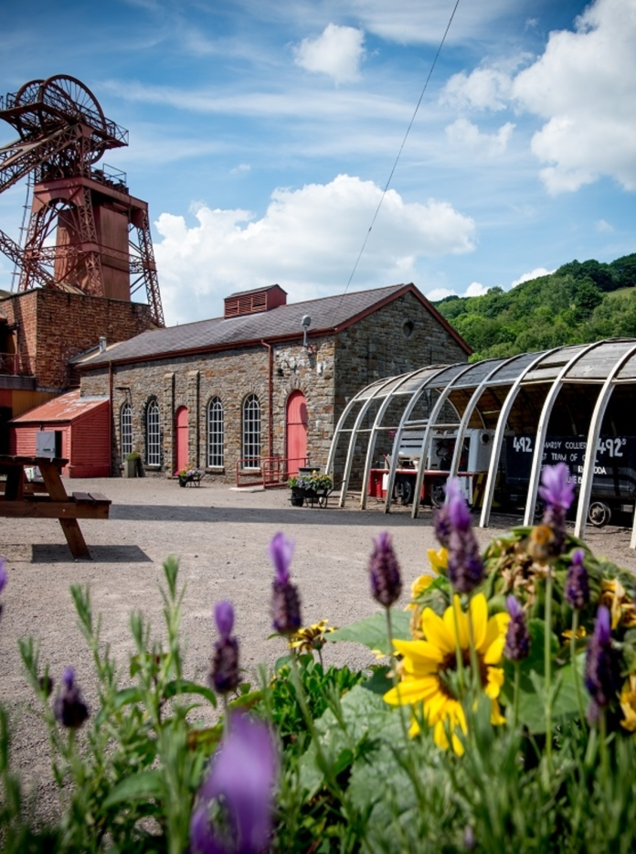 Tu allan i amgueddfa Taith Pyllau Glo Cymru ar ddiwrnod braf gyda blodau ym mlaen y llun.
