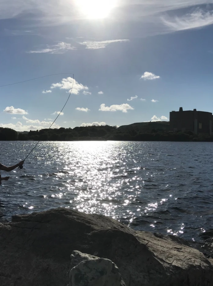 Amlinell rhywun yn sefyll ar lwyfan pysgota, gyda’r llyn y tu ôl iddo, yr haul yn uchel yn yr awyr ac amlinell adeilad tywyll yn y cefndir