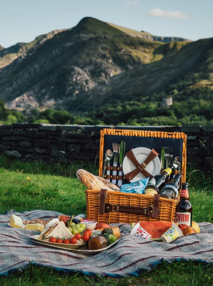 Basged bicnic ar flanced gyda phlatiad o fwyd picnic. Mae mynyddoedd ac awyr las yn y cefndir.