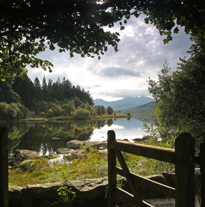 Golygfa o'r llyn o giât gyda mynyddoedd a choed yn adlewyrchu yn y dŵr.