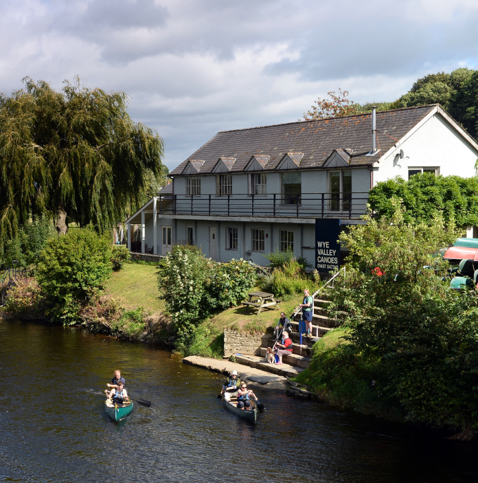 Pobl yn canwio ar yr Afon Gwy gydag adeilad Wye Valley Canoes ar y lan yn y cefndir