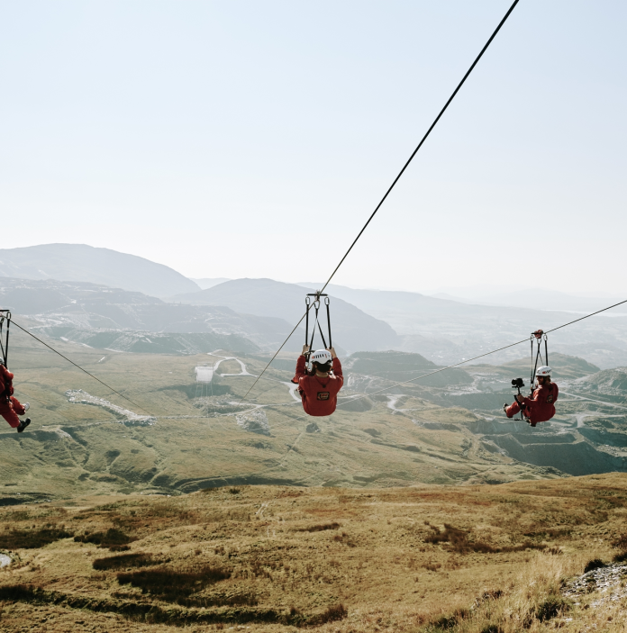 Tri o bobl yn marchogaeth zip-line yn uchel uwchben chwarel lechi.