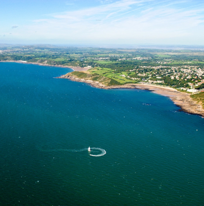 Llun o'r awyr o gwch hwylio ar y môr a Bae Langland yn y pellter