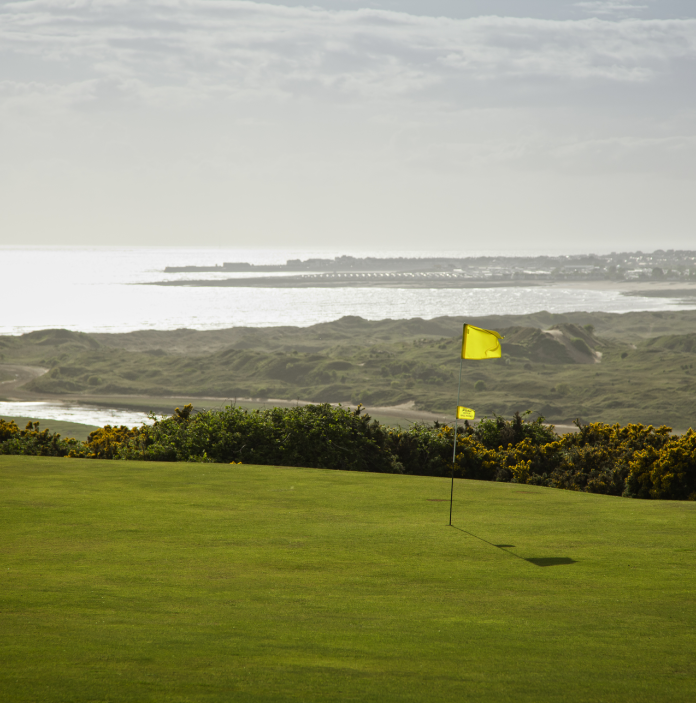 Coes fflag ar y grîn yng Nghwrs Golff Southerndown gyda'r môr a darnau o dir yn y cefndir