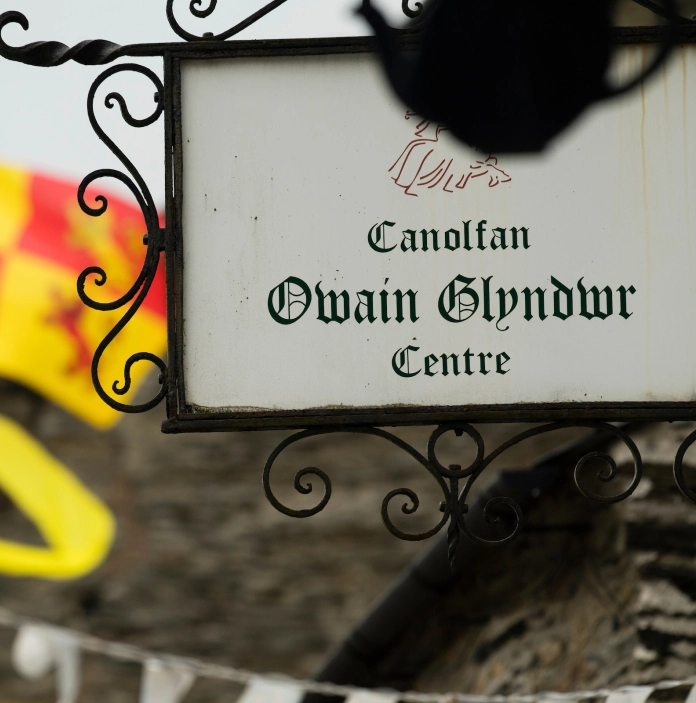 Llun o arwydd Canolfan Owain Glyndŵr, gyda baner Owain Glyndŵr yn chwifio yn y cefndir