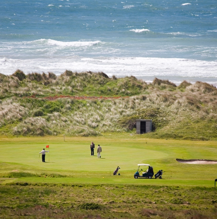 pobl yn chwarae golff ar grîn gyda'r môr yn y cefndir